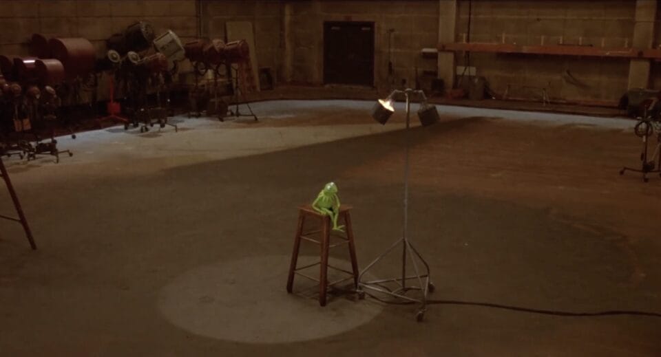 a still from a Muppet film showing Kermit the Frog onstage on a stool