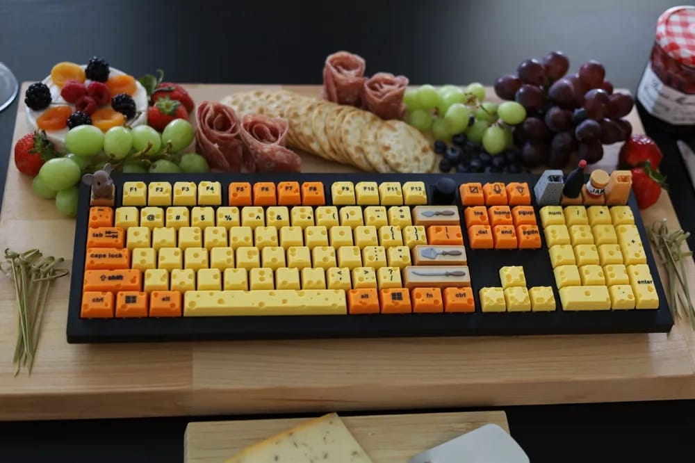 a keyboard with key covers shaped like cheese and a charcuterie board. an actual charcuterie board is behind