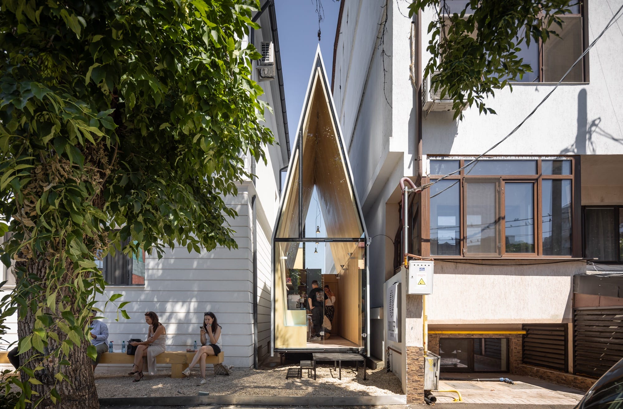 a tiny, contemporary cafe with a steep, pointed roof sits between two larger conventional buildings, designed by Vinklu