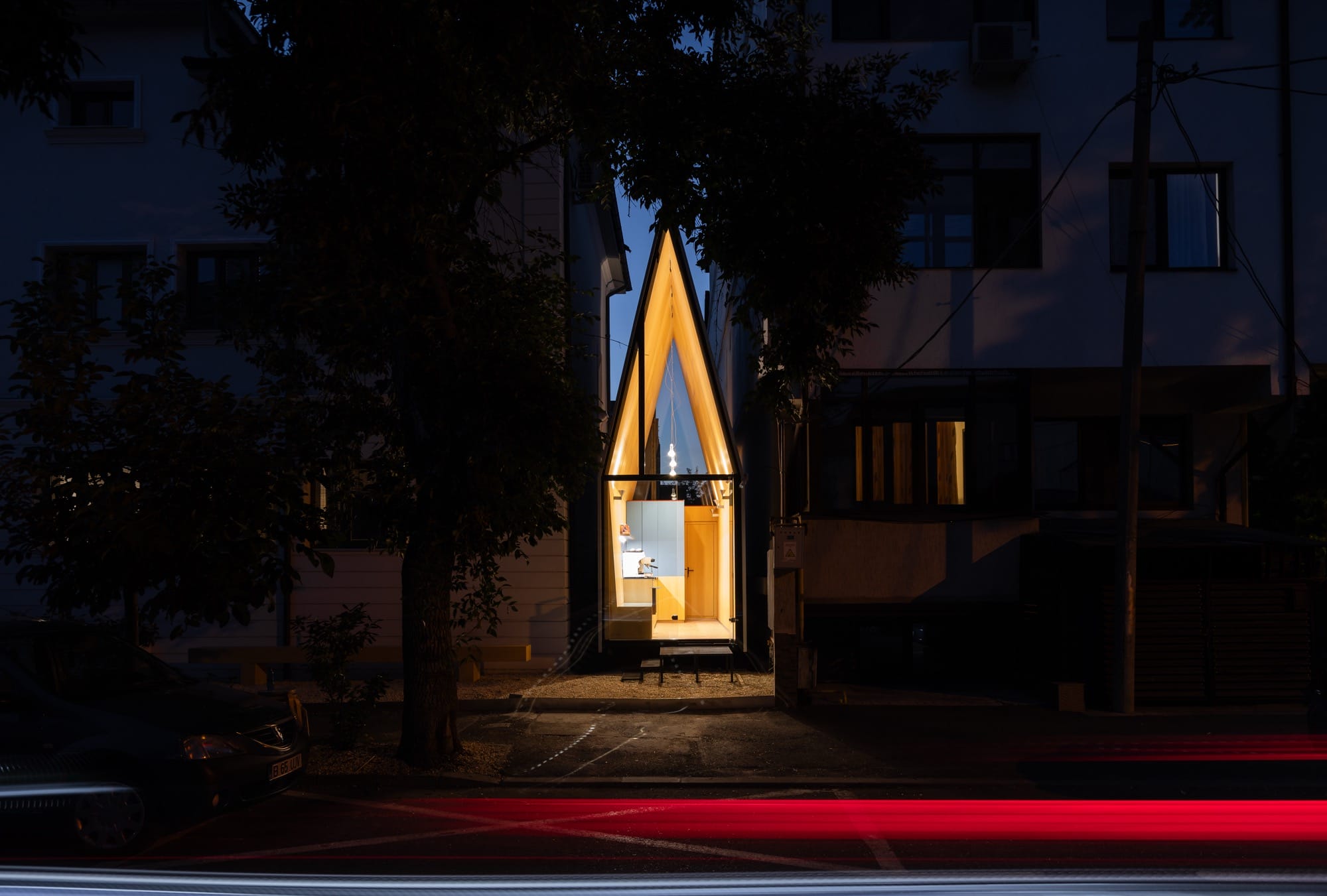a tiny, contemporary cafe with a steep, pointed roof sits between two larger conventional buildings, designed by Vinklu and photographed at night
