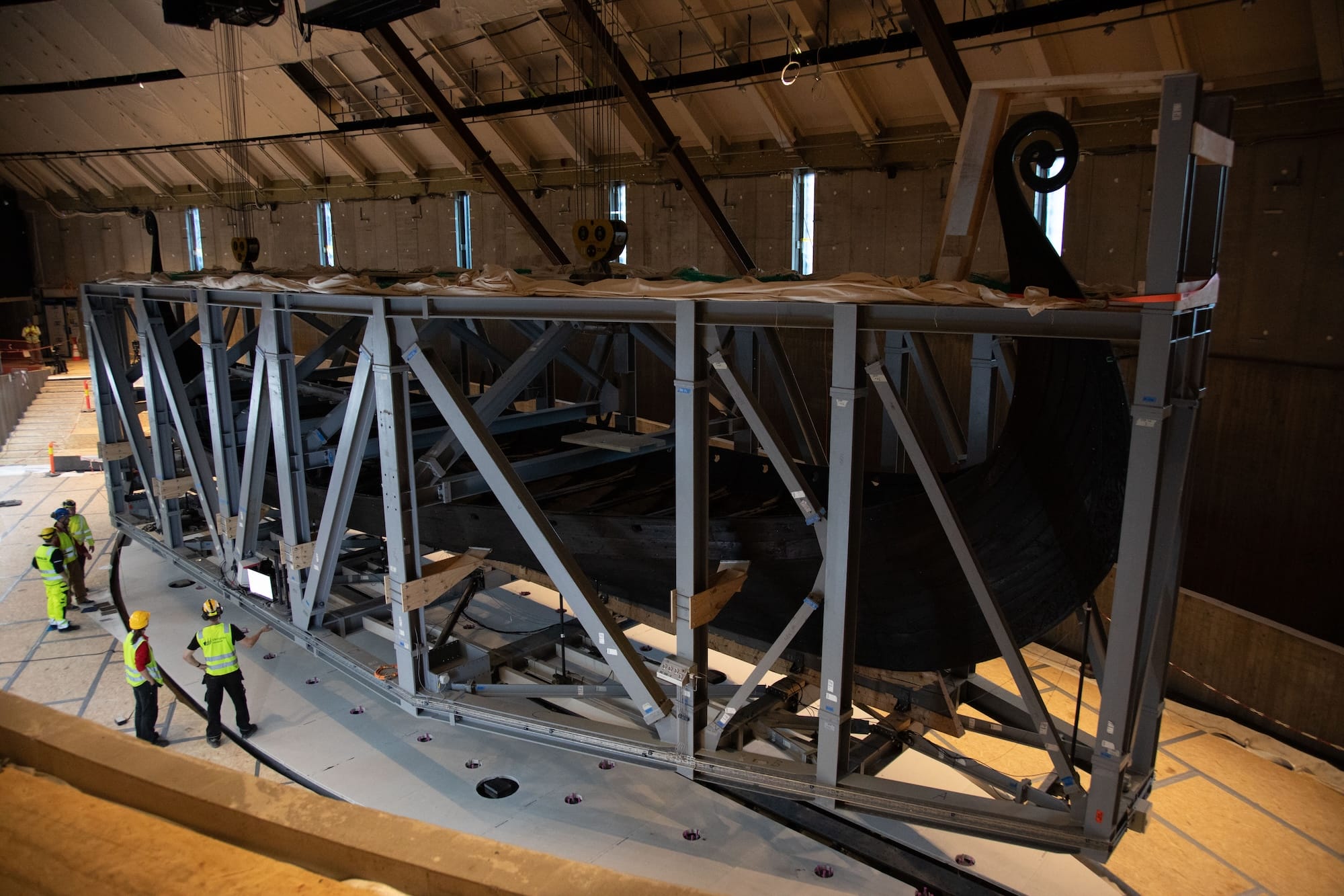workers in hi-vis stand next to the Oseberg Viking Ship as it's moved through a museum in a huge steel crate
