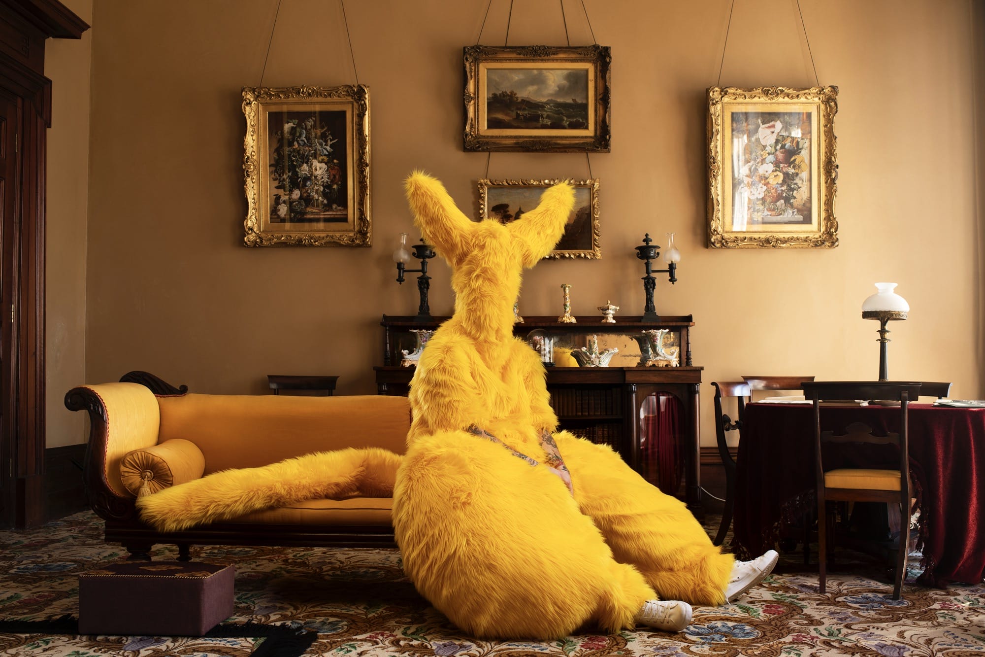 a photograph by Gerwyn Davies of himself in a yellow, mouse-like, full-body costume inside of a room with old paintings on the wall