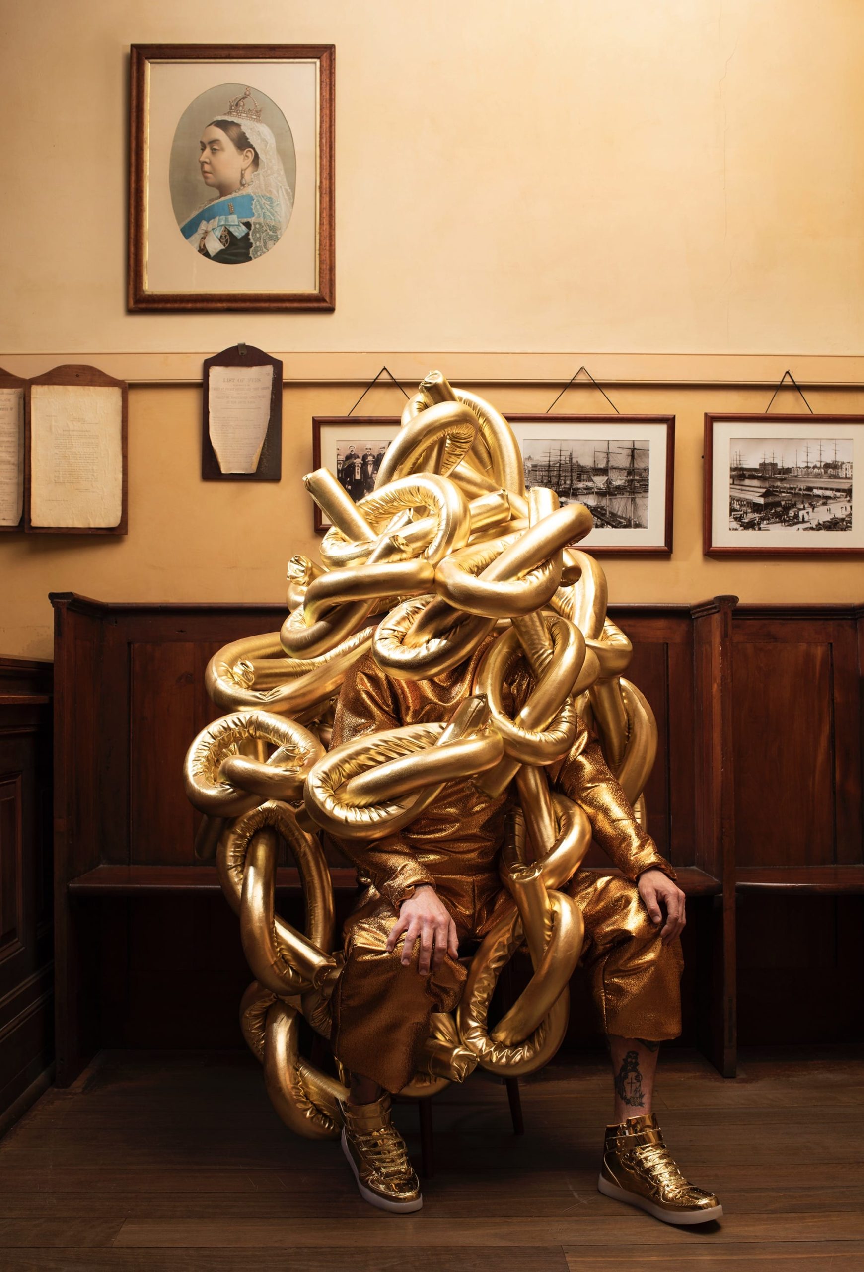 a photograph by Gerwyn Davies of himself in a gold, abstract, full-body vinyl costume inside of a room with old photos on the wall