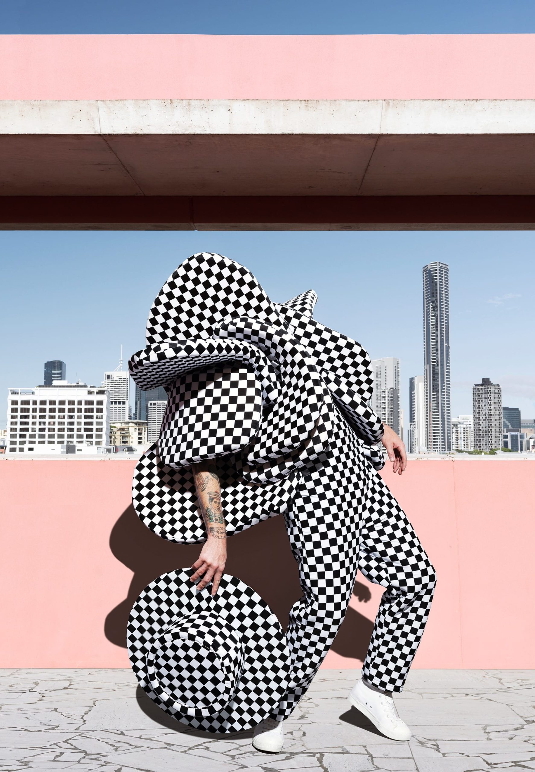 a photograph by Gerwyn Davies of a black-and-white checked, abstract costume obscuring the top half of his body against a city skyline