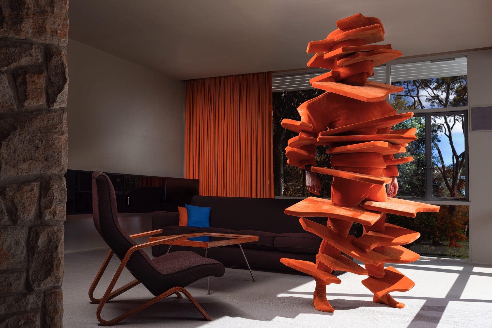 a photograph by Gerwyn Davies of himself in a bright orange, abstract, full-body costume inside of an apartment room